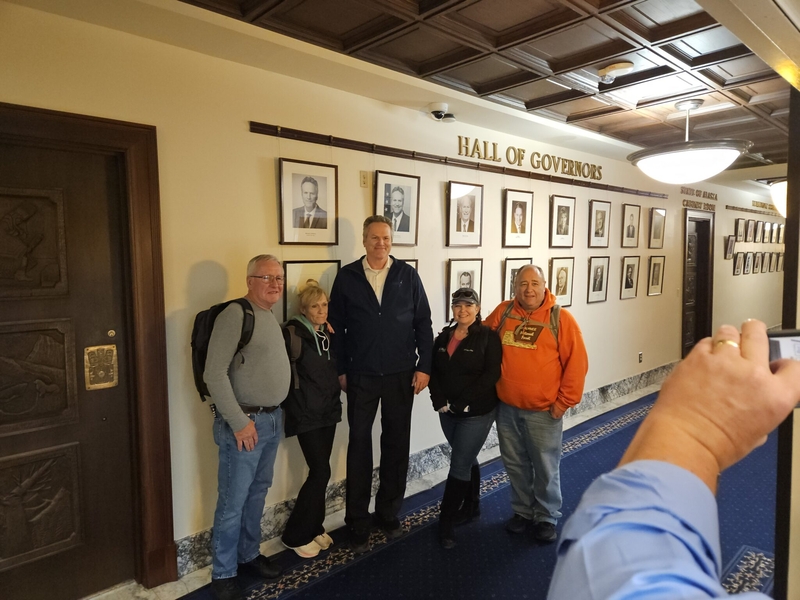Gov. Mike Dunleavy poses for a photo with tourists visiting the Capitol on July 15, 2026. (Photo by Corinne Smith/Alaska Beacon)