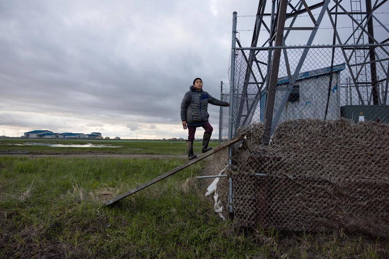 Seth Friend, 12, stands at the base of a wind turbine in Kwigillingok, Alaska, on June 18, 2026. Grass tangled in the fence marks the height reached by floodwaters from ex-Typhoon Halong. (Katie Basile for Anchorage Press)