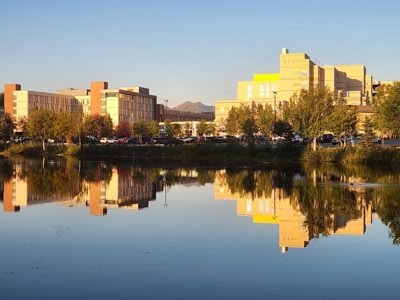 The Alaska Native Medical Center and other buildings on the Alaska Native Tribal Health Consortium campus in Midtown Anchorage are reflected in lake water on Aug. 26, 2026. (Photo by Yereth Rosen/Alaska Beacon)