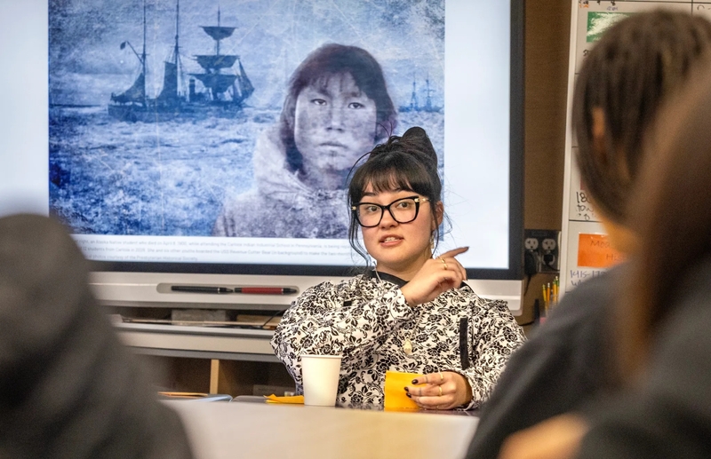 Nyché Andrew talks to a group of students at the Shishmaref School on Oct. 5, 2026, about her journey to bring the remains of her relative, Tummassak, home to Shishmaref, Alaska. The photo collage by Charles Fox on the screen behind her is from an ICT story about the journey of Tummassak, who died more than 100 years ago while attending the Carlisle Indian Industrial School.