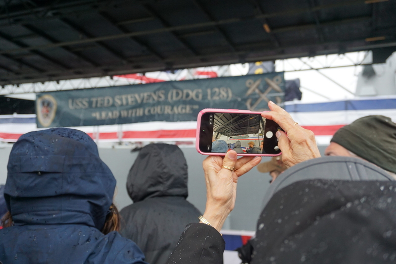 A visitor takes a photo of USS Ted Stevens' banner during an Oct. 1, 2026, media tour of the new Navy warship. (Photo by Yereth Rosen/Alaska Beacon)