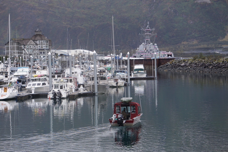 A small recreational vessel sails on Oct. 1, 2026, through the Whittier harbor, where the USS Ted Stevens towers over the docked boats. The new warship is named after the late U.S. Sen. Ted Stevens, a World War II veteran. (Photo by Yereth Rosen/Alaska Beacon)
