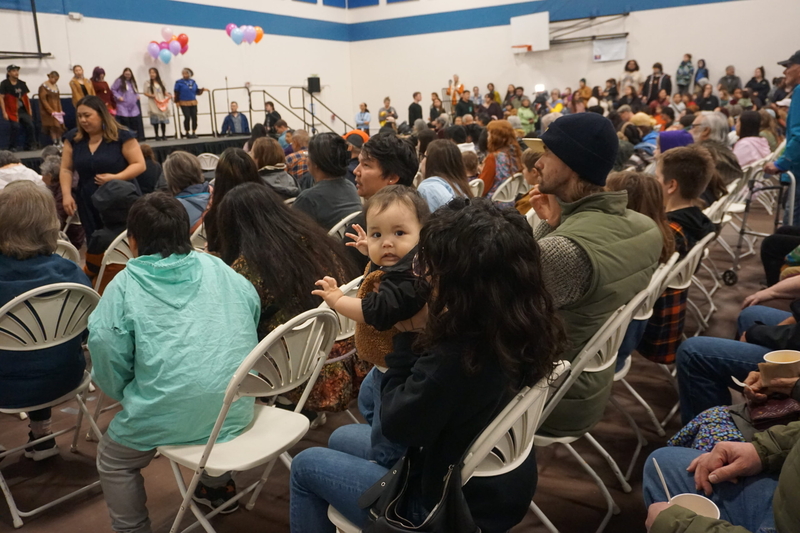 One-year-old Hector Triplett is held by his mother, Hannah, as they watch a dance performance at the Oct. 12, 2026, Indigenous Peoples Day celebration at Alaska Pacific University. (Photo by Yereth Rosen/Alaska Beacon)