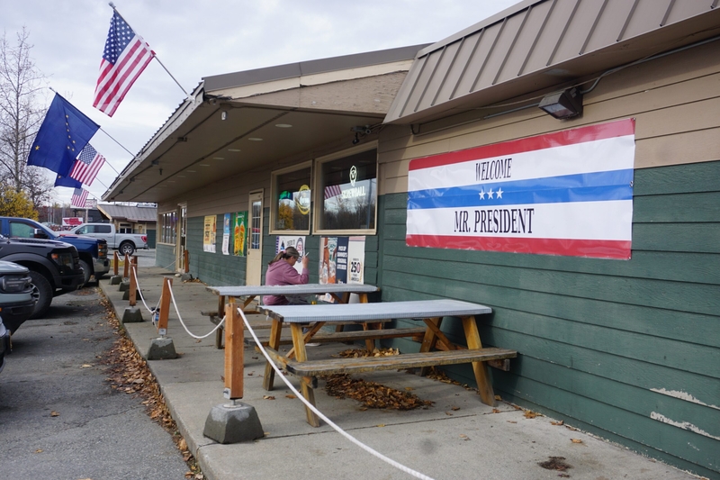 A banner hanging on the outside wall of Tailgaters Sports Bar &amp; Grill in Wasilla on Oct. 13, 2026, welcomes President Donald Trump to city. Trump came to Wasilla to campaign for U.S. Sen. Dan Sullivan, R-Alaska. (Photo by Yereth Rosen/Alaska Beacon)