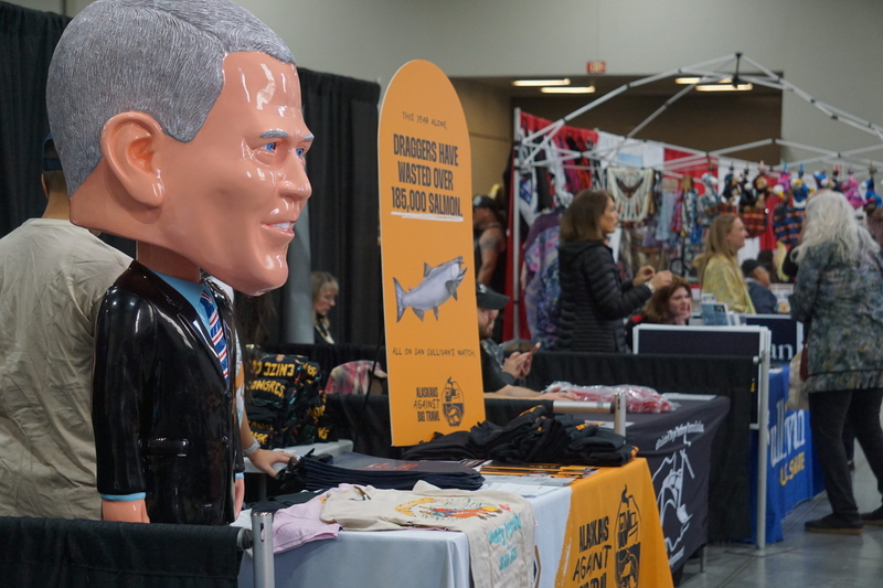 A plastic statue representing U.S. Sen. Dan Sullivan, R-Alaska, labeled "Yes Man Dan," stands on Oct. 23, 2026, at a booth manned by the liberal-leaning 907 Initiative at the Alaska Federation of Natives convention arts fair. The "Yes Man Dan" slogan is part of the group's criticism of Sullivan for his allegiance to President Donald Trump. The booth also features banners criticizing Sullivan on fishery management issues. Nearby is a booth promoting Sullivan's reelection and manned by the senator's wife, Jul