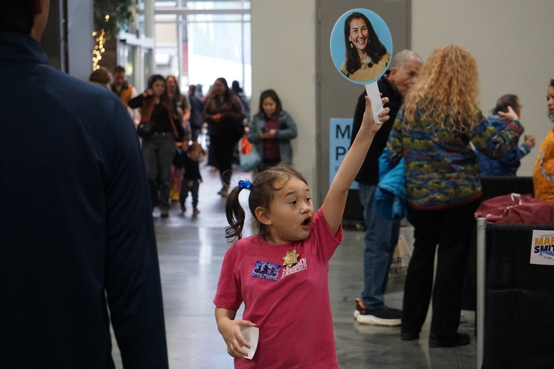 A young supporter of Mary Peltola holds up a paddle bearing the Democratic candidate's image as she walks on Oct. 24. 2026, through the arts fair that is part of the Alaska Federation of Natives annual convention. Peltola, who is Yup'ik, is challenging incumbent Republican Sen. Dan Sullivan. (Photo by Yereth Rosen/Alaska Beacon)