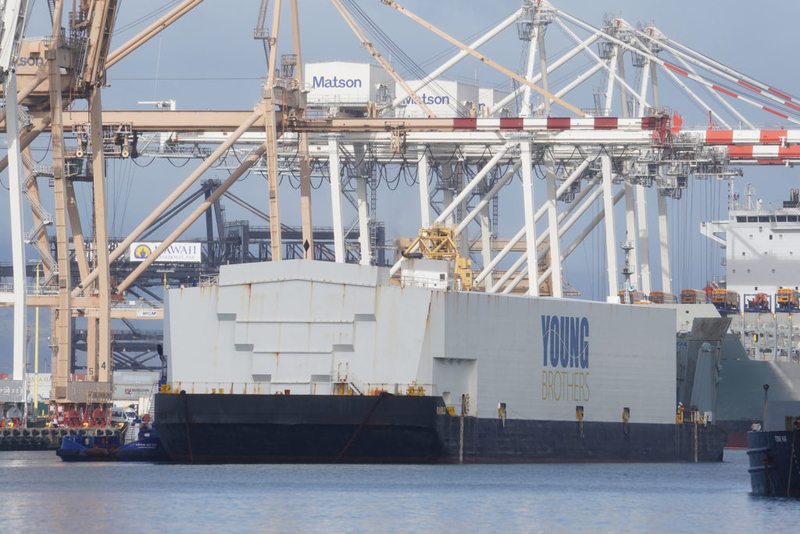 Tugboats guide a large Young Brothers barge inside Honolulu Harbor with Matson cranes in the background.