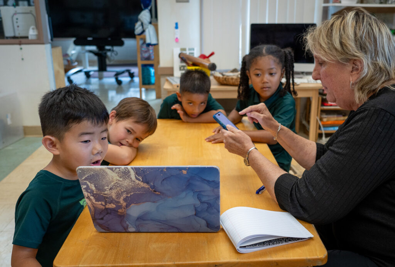 Four children wearing dark green t-shirts sit at a table looking at a laptop screen with a woman holding a mobile phone.