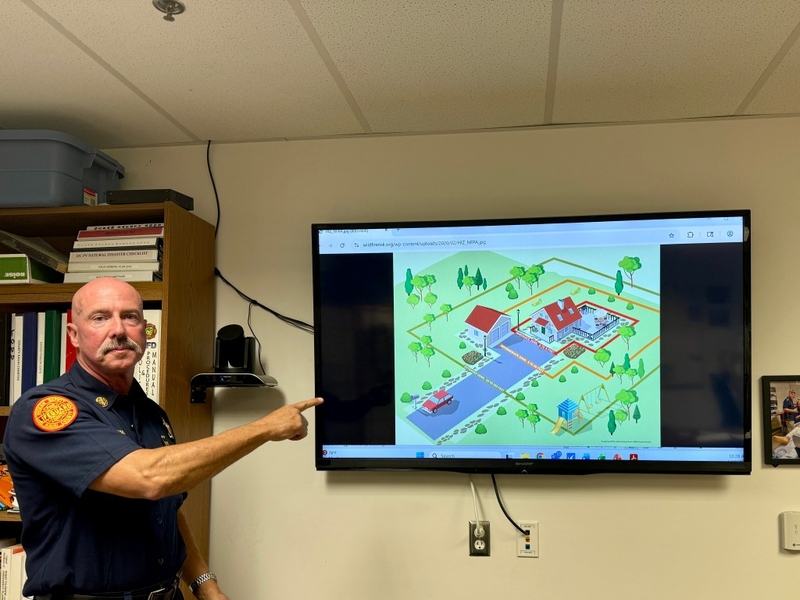 A balding man with a gray mustache points at a television screen that depicts a home and yard with boundaries close to and away from the house.