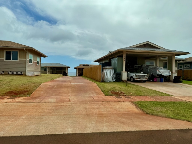 Two cars sit in an open carport. To the left, a driveway leads to two more houses in the background.
