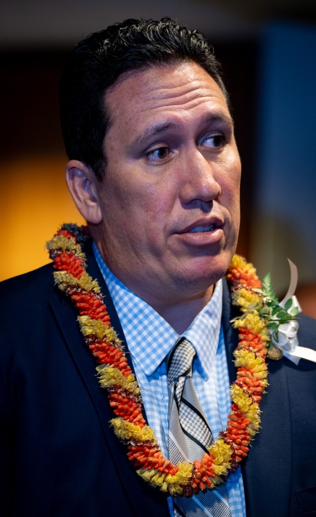 A head and shoulders photo of a man with short dark hair. He wears a checked blue shirt, diagonal gray tie, blue blazer and a yellow and orange lei.