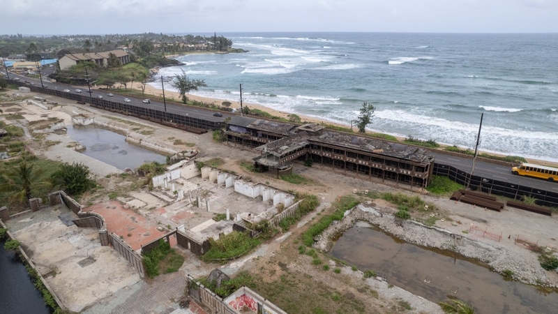 A long dark mostly demolished building sits next to a highway with the ocean in the background. In the foreground is standing water and debris.
