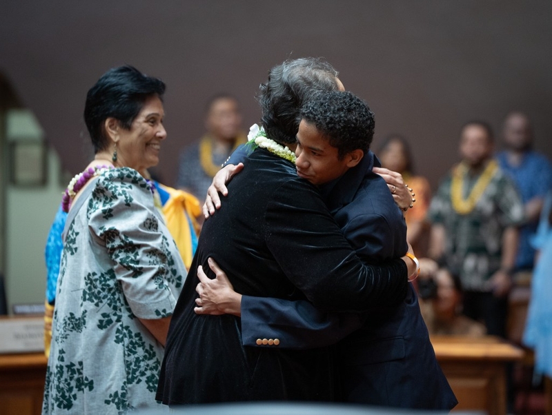 A young man wearing a blue blazer hugs a woman wearing a black blazer.
