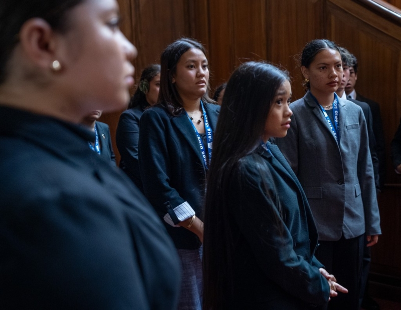 Young women dressed in blazers look to the right of the camera as they stand and listen to instruction.