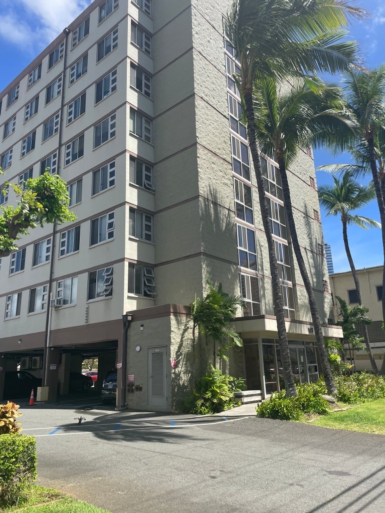 Grey eight-story apartment building with palm trees in front of it.