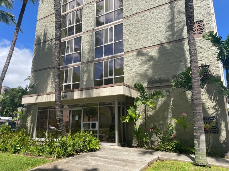 A greenish grey apartment building photographed from the street, with several palm trees in the foreground.