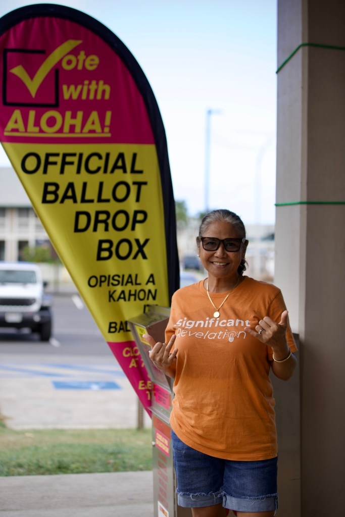 Angelina Abapo, wearing an orange t-shirt and her hair in a ponytail, smiles and stands in front of the official voting dropbox in front of the Velma McWayne Santos Community Center.