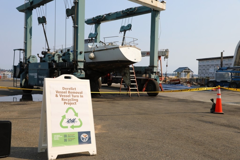 Yacht suspended in a heavy lifter in the background. A sandwich board in the foreground explains a program for voluntarily turning in derelict vessels.