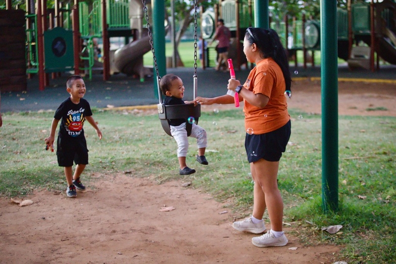 A woman pushes a young child on the swing as another child smiles and watches. Bubbles float by.