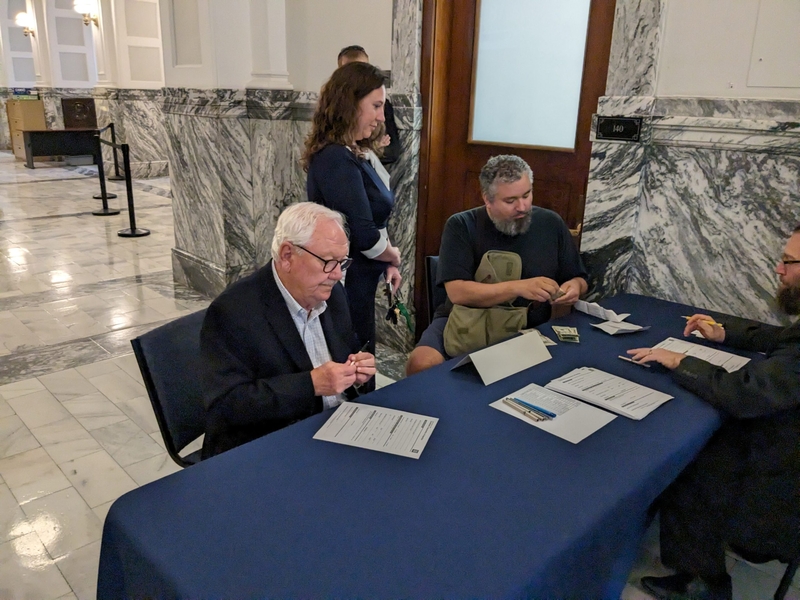 McPherson Sen. Rick Wilburn, left, signs papers at the Kansas secretary of state's office to be a candidate for the Kansas House seat vacated by a medical emergency forcing McPherson Rep. Les Mason to withdraw. To Wilburn's left is Eddied Martinez, a Lawrence Democrat, who filed to run in the 42nd District of the Kansas House. (Tim Carpenter/Kansas Reflector)