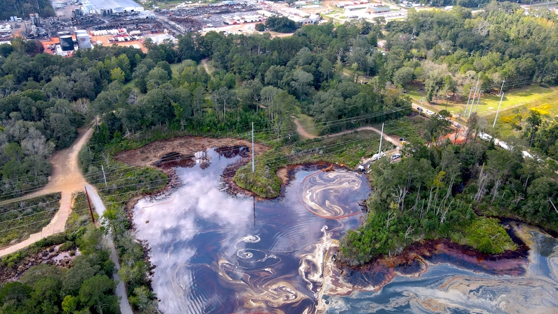 Drone view of swirls of oil and chemicals covering water surface with destroyed Smitty's Supply plant in background
