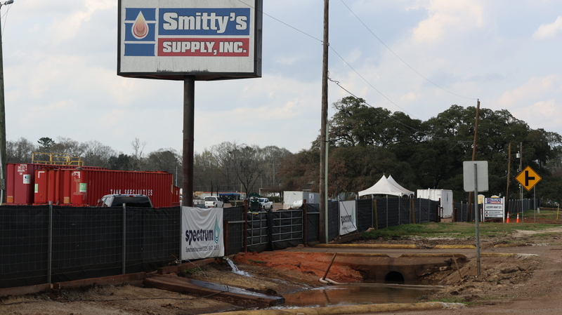 Water gushes into messy ditch from large hose with "Spectrum" banner hangs on fence directly over hose; Smitty's plant in background