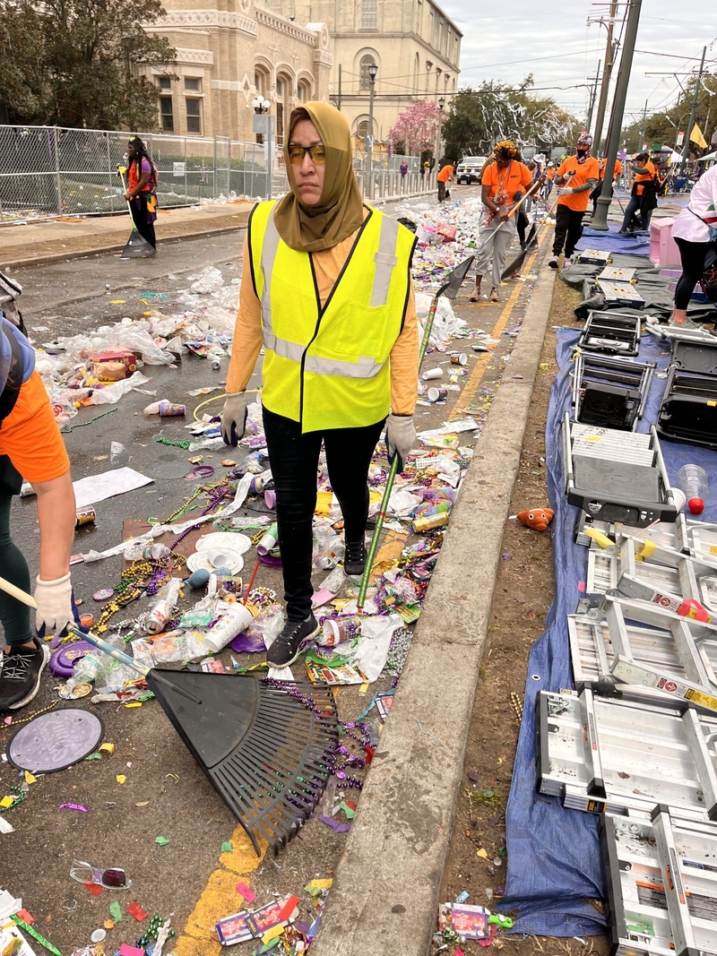 A clean-up worker walks amid debris from a Mardi Gras parade along St. Charles Avenue as another worker rakes away the mess.