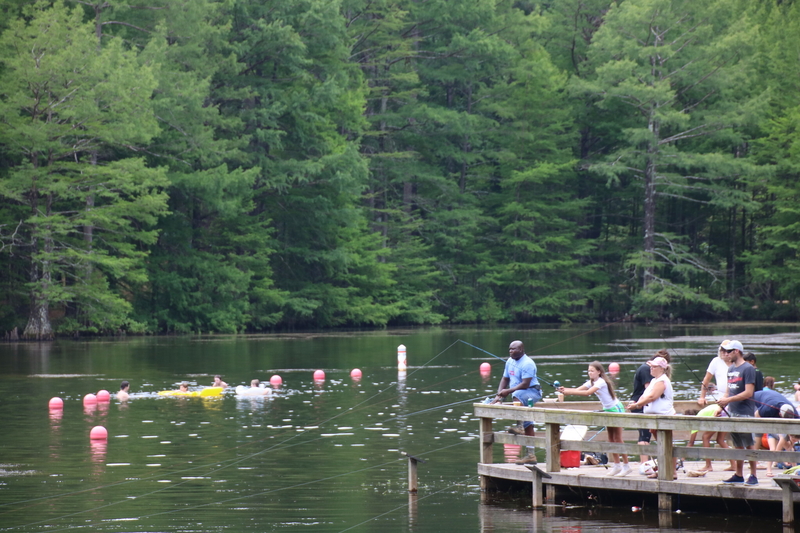 People swim in Stuart Lake in Kisatchie National Forest as people holding fishing polls while standing on a pier extending into the lake.