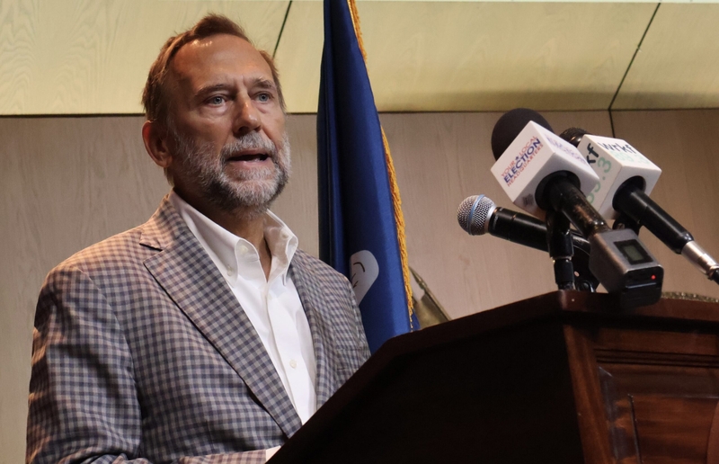 Pat Forbes stands behind a podium with microphones places on its edge.