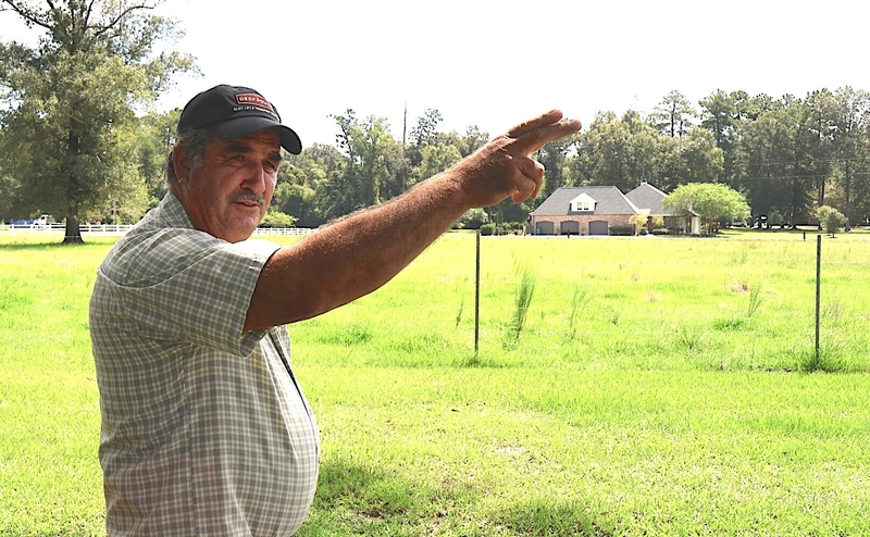 Man in a hat and plaid shirt stands in front of a field and gestures with his hands.