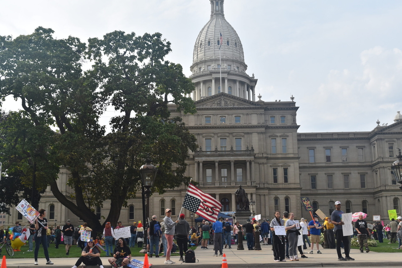 A woman holds an upside down American flag that reads 'SOS' in front of the Michigan State Capitol.