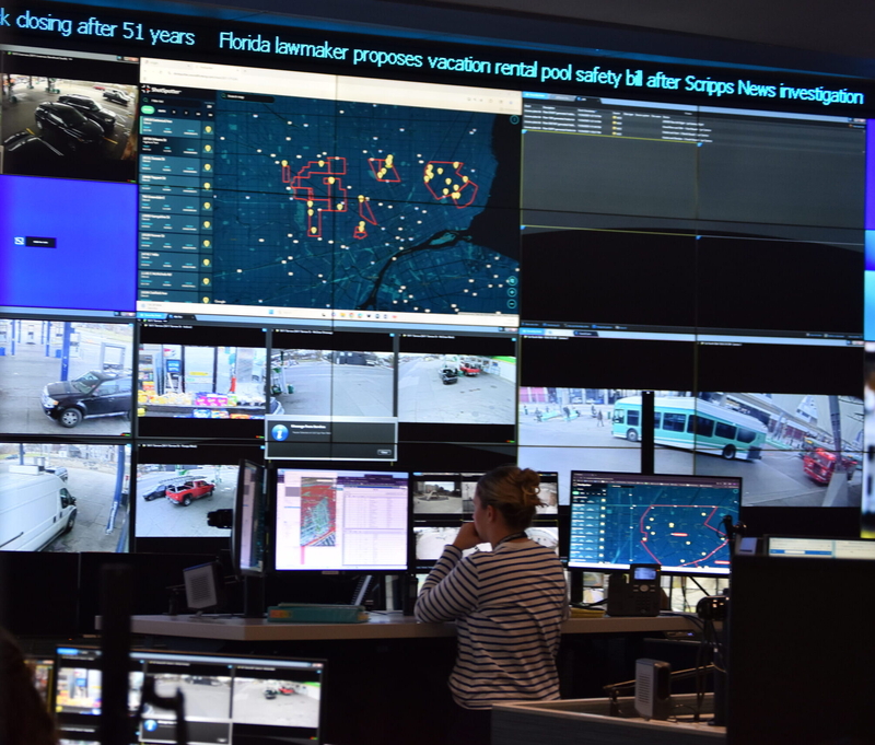 A woman sits at computer screens, with a wall of larger screens, including a map with pinpoints of ShotSpotter alerts, in front of her.