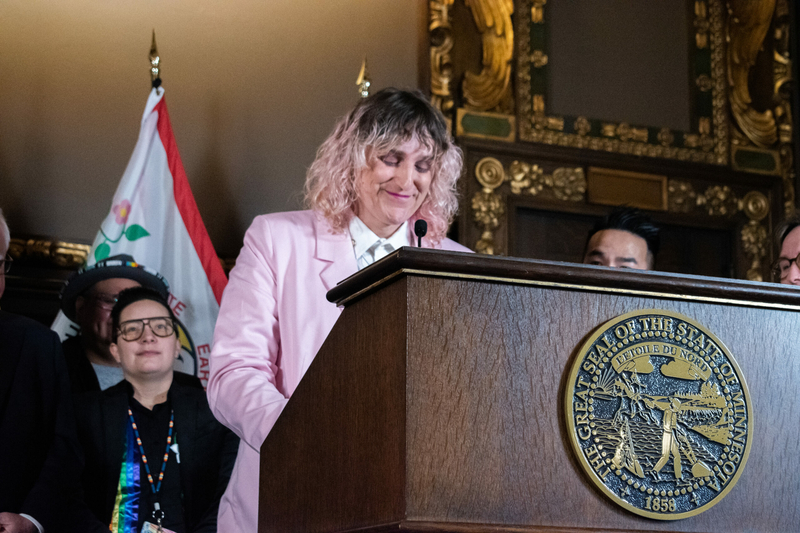 A woman with dark roots and pastel pink hair in a pastel pink suit stands behind a podium and looks down, with a slight smile on her face.