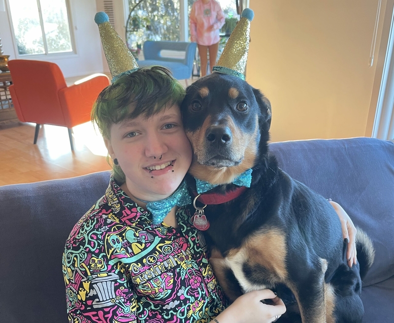 A person wearing a colorful graffiti shirt with green hair hugs a black and brown medium-sized dog in a bowtie. Both are wearing little party hats.