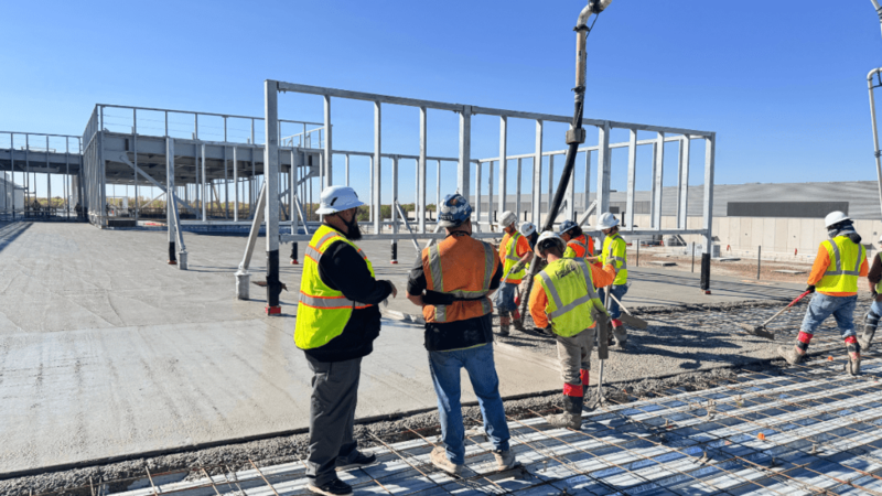 A crew of construction workers in hard hats and high-visibility vests pour concrete from an overhead pump onto a rebar-reinforced deck, with steel framing for a building rising in the background under a blue sky.