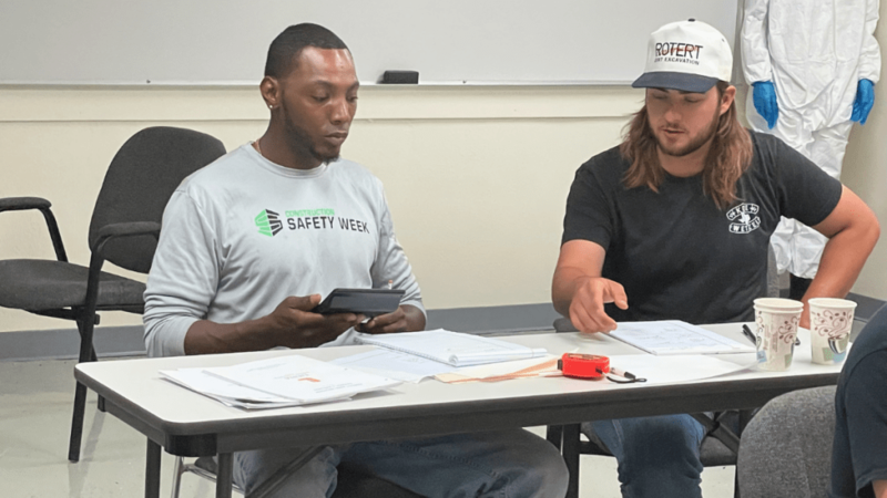 Two young men sit at a folding table reviewing paperwork and using a calculator during a construction trades classroom session, with notebooks and a tape measure spread across the table.