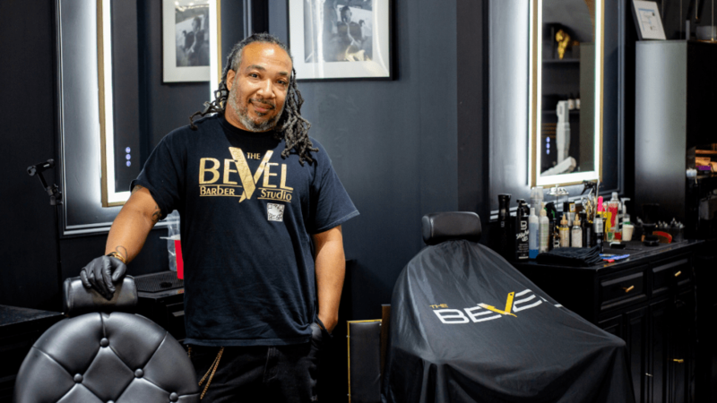 Casie Murff, a barber with long locs and a gray-flecked beard, stands smiling in a black