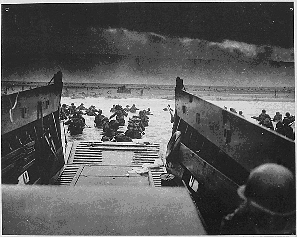 Down the ramp of a Coast Guard Landing barge Yankee soldiers storm toward the beach-sweeping fire of Nazi defenders in the D-Day Invasion of the French Coast. Troops ahead may be seen lying flat under the deadly machine gun resistance of the Germans. Soon the Nazis were driven back under the overwhelming Invasion forces thrown in from Coast Guard and Navy amphibious craft. (National Archives)