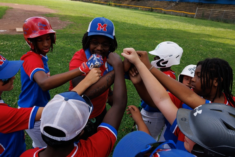 A youth baseball team in red and blue uniforms gathers in a huddle on the field, joining hands in the center for a team cheer.
