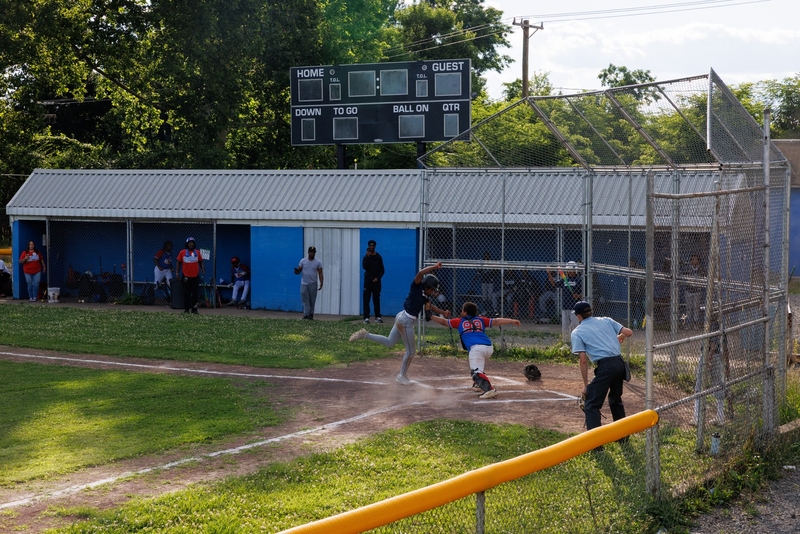 A baseball player runs to home plate as the catcher prepares to tag, with the umpire observing and team members watching from the dugout.