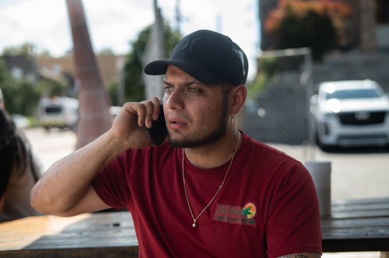 A man in a red shirt and black cap sits outdoors at a table, holding a phone to his ear and looking to the side.