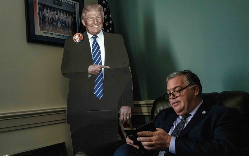 A stout man with gray hair and glasses sits next to a cut out figure of President Donald Trump.