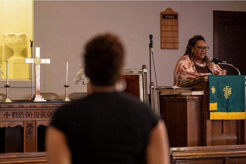 A Black woman stands at the front of a church and appears to be preaching.