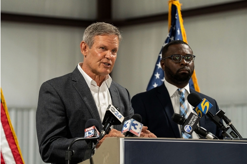 A white man speaks into a microphone as a Black man next to him looks out the side of his eyes.