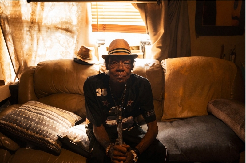 A man wearing a hat and holding a cane sits on a sofa and looks at the camera.