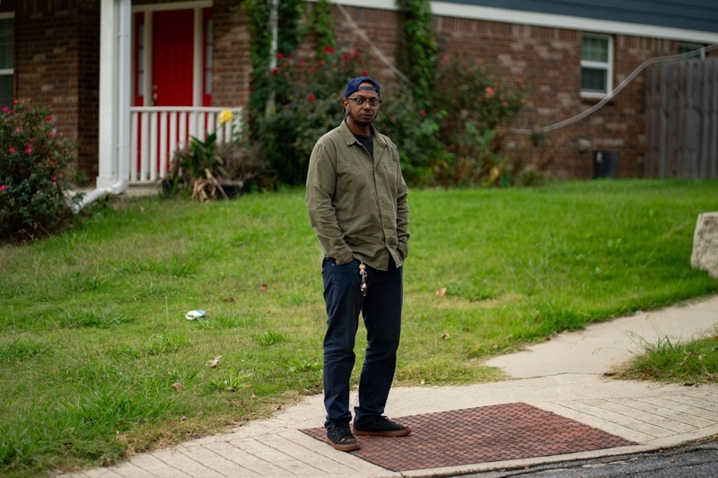 A Black man with his hands in his pockets stands in front of a brick house with a lawn.