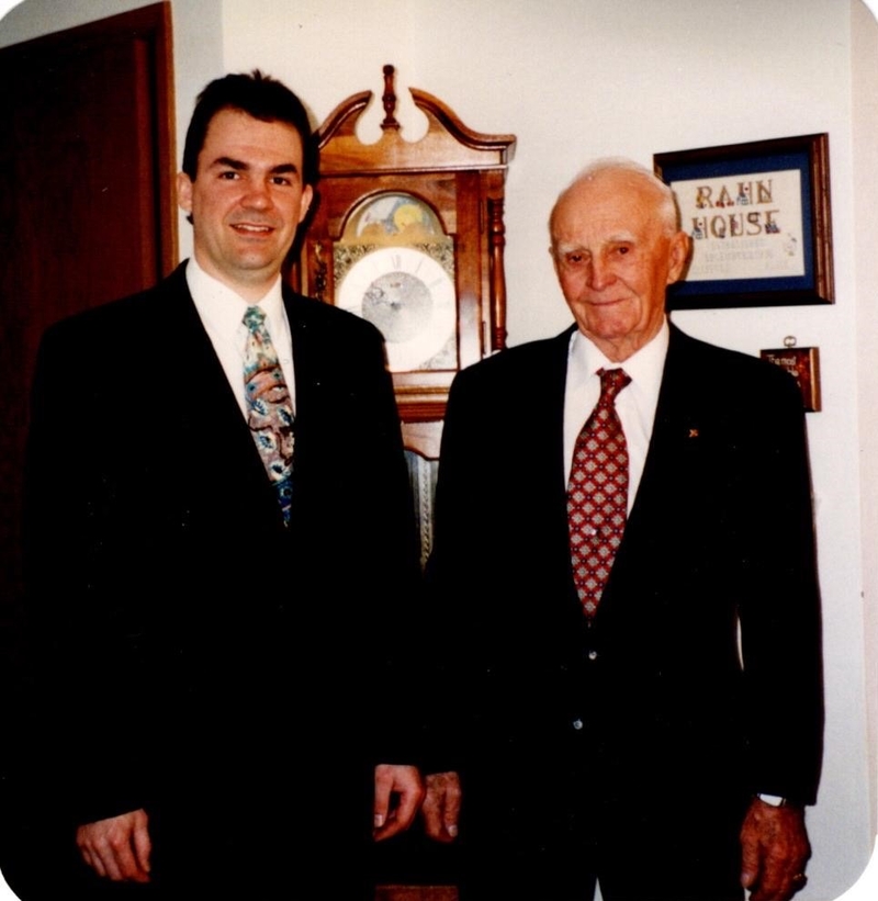 An older man with thinning white hair stands next to a younger, taller man with dark hair. Both wear dark suits.